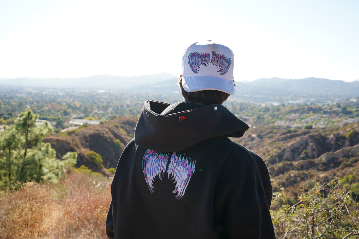 Winged Always Prosper back print on The Skelly hoodie, worn on a hilltop above the valley
