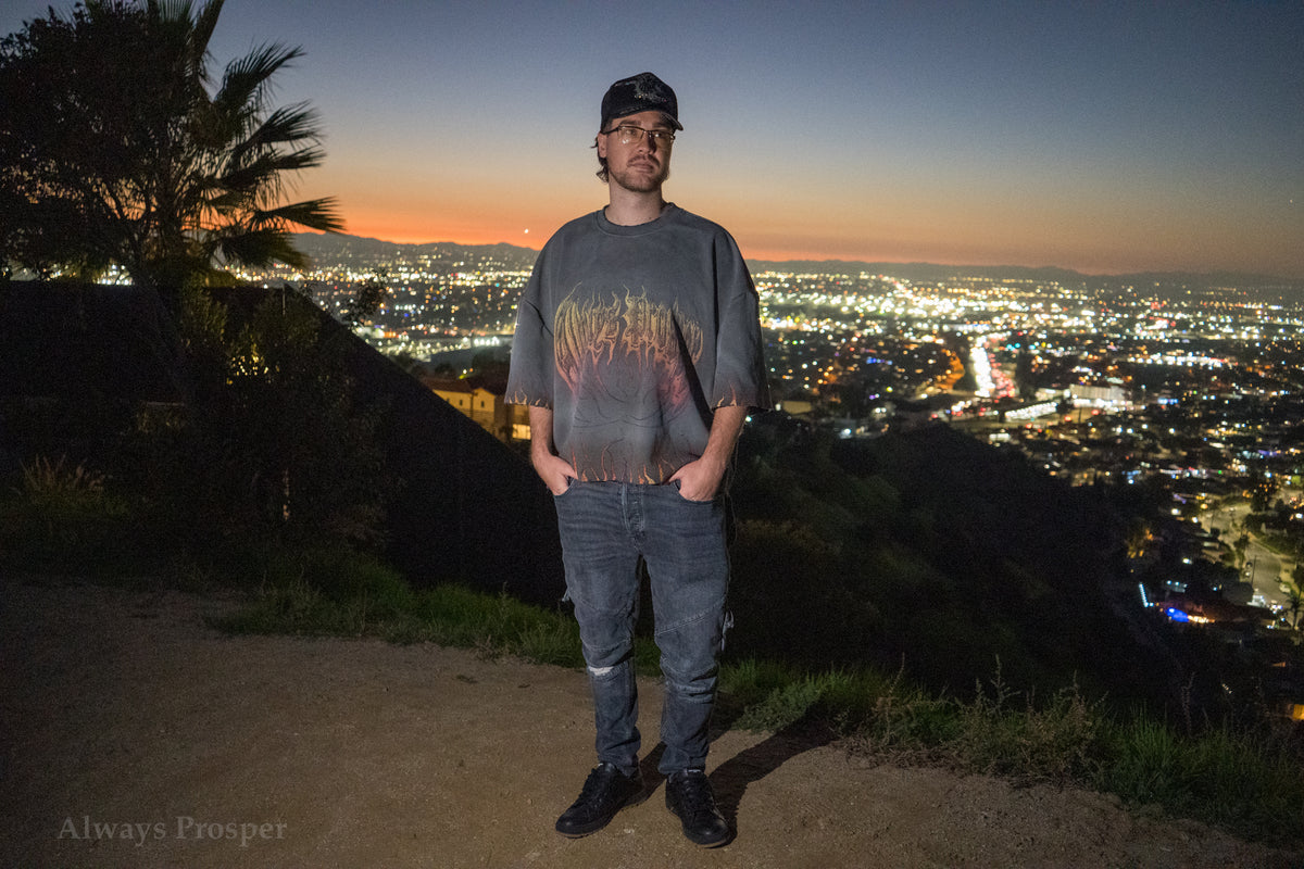 Model wearing the Always Prosper Through the Flames tee on a hillside above the city at sunset