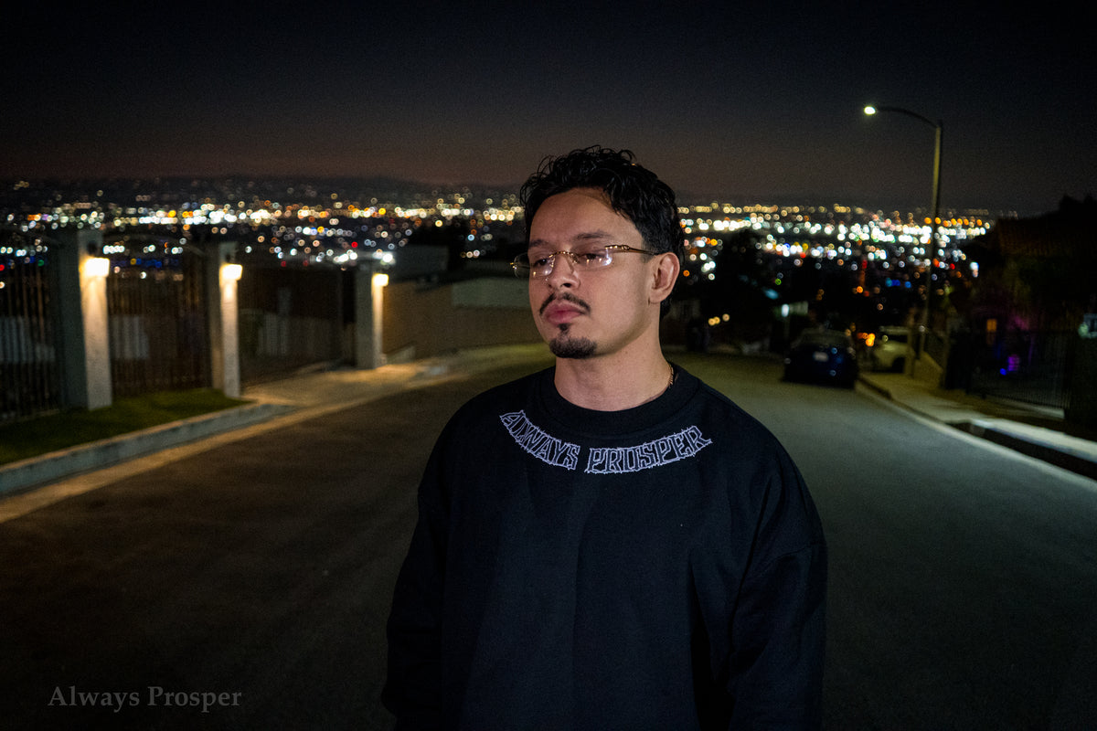 Model wearing the By Any Means sweater on a hilltop street at night, collar print visible