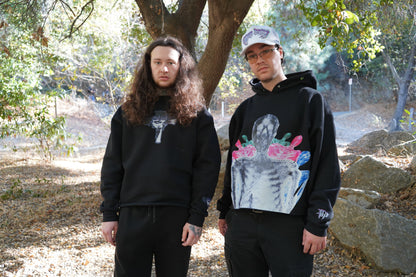 Two models outdoors, one in the Always Prosper Trinity crewneck and one in The Skelly hoodie